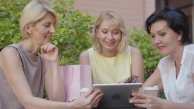 Açık kafede tablet kullanan üç orta yaşlı kadın. İnternette komedi ya da stand-up şovu izleyen neşeli beyaz kadın portresi. Mutlu arkadaşlar sosyal medyayı kullanır ve açık havada gülerler..