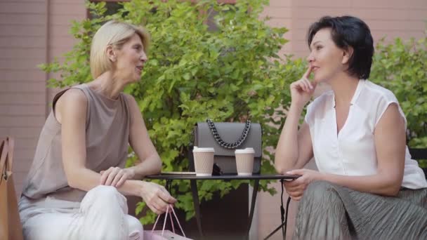 ブルネットの友人にピンクのショッピングバッグで贈り物を与える肯定的な中大人の女性の側面図。2人の幸せな白人女性の肖像画屋外カフェで休憩し、話しています。女性の友情. — ストック動画