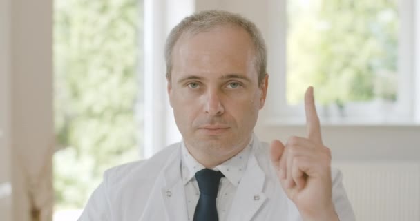 Retrato de primer plano del médico serio y confiado sentado con el dedo levantado y mirando la cámara. Médico profesional caucásico de mediana edad posando en el hospital. Sede del cine 4k ProRes. — Vídeos de Stock