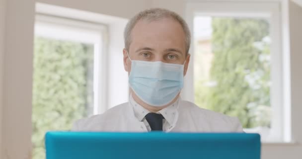 Retrato de médico caucásico serio en mascarilla que habla en la cámara selfie en el ordenador portátil y el gesto. Médico varón de mediana edad que consulta a personas en línea sobre el bloqueo del coronavirus. Sede del cine 4k ProRes. — Vídeos de Stock