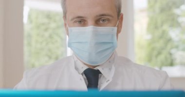 Laptop Selfie makinesinde konuşan maskeli orta yaşlı doktorların yakın çekimi. Sanal konferansta Covid-19 salgınını tartışan kendine güvenen beyaz bir adamın portresi. Sinema 4k ProRes Merkezi.