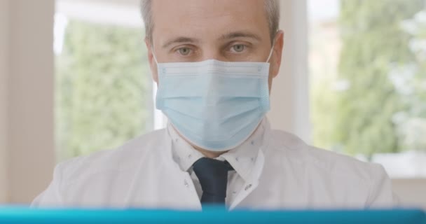 Primer plano del médico de mediana edad en la máscara de la cara hablando en la cámara selfie portátil. Retrato de un hombre caucásico serio y confiado discutiendo la pandemia de Covid-19 en una conferencia virtual. Sede del cine 4k ProRes. — Vídeos de Stock