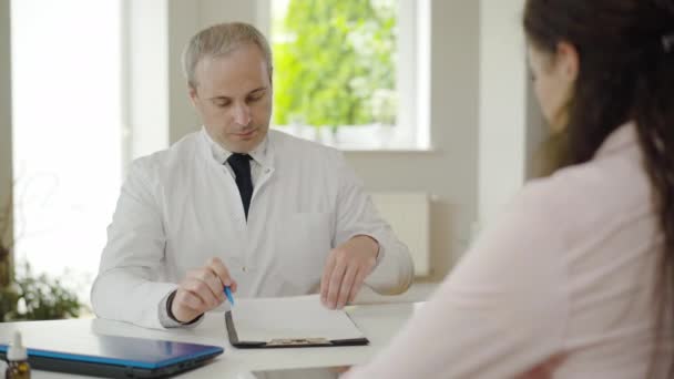 Retrato de un médico serio de mediana edad que revisa los resultados del examen de los pacientes y agita la cabeza. Médico caucásico consultando a mujer enferma en el hospital. Concepto de medicina y asistencia sanitaria. — Vídeo de stock