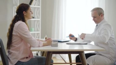 Genç, beyaz bir kadının doktorla birlikte hastanede oturup konsültasyonu dinlemesinin yan görüntüsü. Klinikte hasta bir hastaya danışmanlık yapan profesyonel bir orta yaşlı adam. Tıbbi konsept.