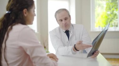 Ciğerlerinde röntgen olan kendinden emin bir doktorun hastayla konuştuğu portre. Önde oturan bulanık genç bayana radyografiyi açıklayan profesyonel bir beyaz adam. Pulmonolog danışma istemcisi.