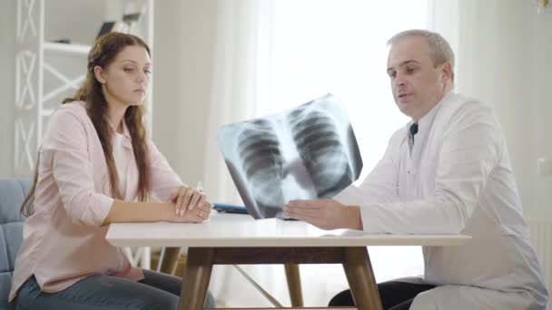 Médico profissional que explica a radiografia dos pulmões a uma jovem no hospital. Retrato de visão lateral do homem adulto médio e da mulher jovem discutindo a condição de saúde dentro de casa. Consulta médica. — Vídeo de Stock