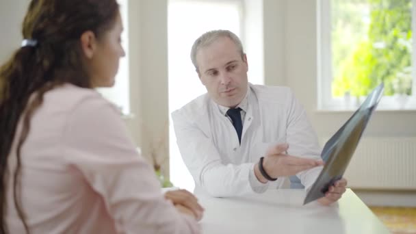 Retrato del doctor confiado hablando con el paciente mostrando radiografía de pulmones. Hombre caucásico profesional explicando la radiografía a una joven borrosa sentada al frente. Cliente consultor de neumología. — Vídeos de Stock