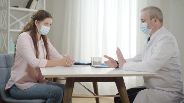 Widok z boku lekarza i pacjenta w maskach siedzących przy stole i mówiących. Wizyta lekarska przy zamknięciu pandemii Covid-19. Kaukaski mężczyzna konsultant kobieta w szpitalu na coronavirus kwarantanny. — Wideo stockowe