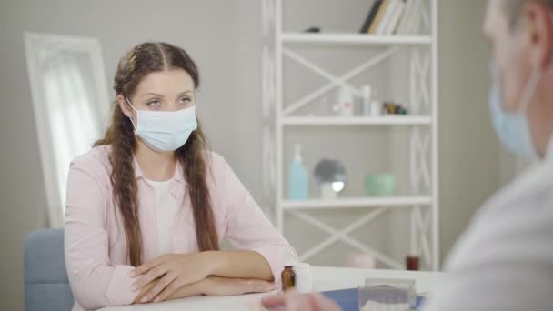 Retrato de una hermosa mujer en máscara facial escuchando al médico en el hospital. Paciente caucásico joven grave sometido a consulta en consultorio médico en cuarentena pandémica de Covid-19. — Vídeos de Stock