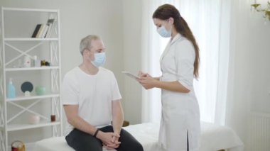 Maskeli doktor Covid-19 salgınında orta yaşlı bir adamın tıbbi geçmişini alıyor. Hasta belirtilerini toplayan ve tablet kullanan profesyonel bir kadın. Coronavirüs tedavisi.