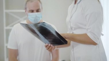 Akciğer röntgeni, tanınmayan bir doktorla birlikte yüz maskesi takan orta yaşlı bir adama röntgen çekiliyor. Profesyonel beyaz kadın doktor hastaya Covid-19 komplikasyonlarını açıklıyor..