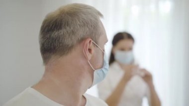 Şırıngayı doldurup kameraya dönen yüzü maskeli korkmuş erkek hastaya yakından bakın. Arka planda ilaç hazırlayan bulanık bir doktorla korkmuş beyaz bir adamın portresi..