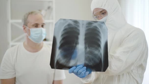 Verschwommener Arzt im antiviralen Anzug im Gespräch mit einem erwachsenen Mann mit Gesichtsmaske, der Covid-19-Komplikationen an Lungenröntgen zeigt. Konzentriert auf Röntgenaufnahmen in den Händen der Ärzte. Coronavirus-Pandemiekonzept. — Stockvideo