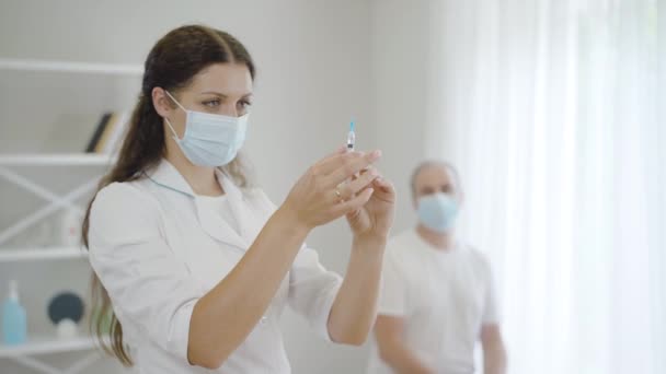 Серйозна красива жінка в масці для обличчя готує ін'єкції для пацієнта, який сидить у лікарні на задньому плані. Портрет впевненої жінки-лікаря або медсестри з вакциною проти ковіда-19 в шприці . — стокове відео