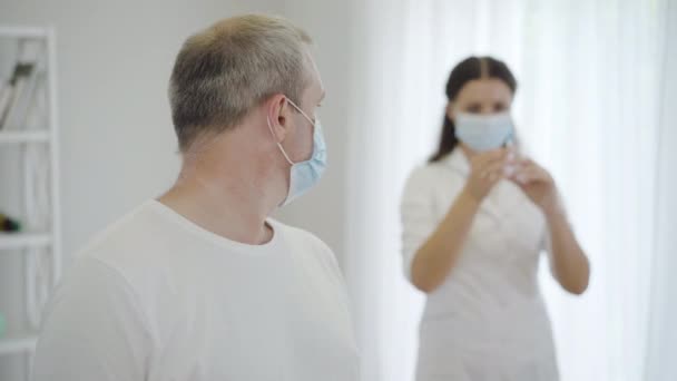 Porträt eines verängstigten erwachsenen Mannes mit Gesichtsmaske, der auf den Arzt mit der Spritze blickt und sich der Kamera zuwendet. Verängstigter kaukasischer Patient wartet auf Injektion gegen Covid-19-Virus durch Krankenschwester. — Stockvideo