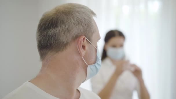 Primer plano del paciente varón asustado con máscara facial mirando hacia atrás a la enfermera llenando la jeringa y girando hacia la cámara. Retrato de hombre caucásico enfermo asustado con médico borroso preparando droga en el fondo. — Vídeos de Stock