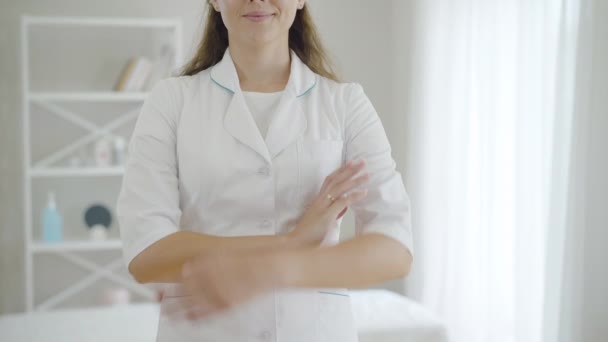 Unerkennbare junge Ärztin, die sich die Hände kreuzt und lächelt. Selbstbewusste brünette Kaukasierin posiert am Arbeitsplatz im Krankenhaus. Hausarzt. — Stockvideo