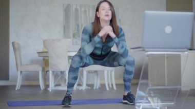 Kendine güvenen güzel bir sporcunun portresi çömelmiş ve dizüstü bilgisayara bakıyor. Genç Kafkas esmer kadın çevrimiçi eğitim izliyor. Covid-19 'daki bireysel eğitmenle kablosuz çalışma.