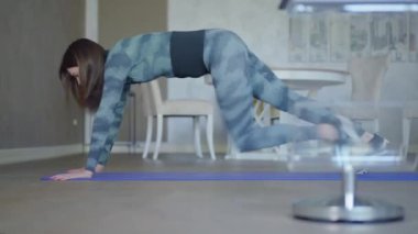 Geniş açılı genç sporcu kadın kapalı alanda kalas egzersizi yapıyor. Evde ya da spor salonunda kendine güvenen beyaz kadın eğitiminin yan görüntüsü. Spor konsepti.