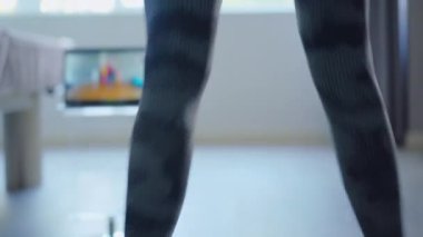 Arka planda bulanık dizüstü bilgisayar ekranı ile atlayan tanınmamış ince sportmen kadının arka görüntüsü. Kişisel antrenör, Covid-19 tecridindeki müşteriye çevrimiçi egzersiz gösteriyor. Coronavirus sporu.