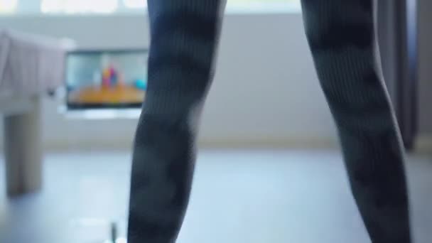 Zadní pohled na nepoznatelnou štíhlou sportovkyni skákající s rozmazanou obrazovkou notebooku na pozadí. Osobní trenér ukazuje cvičení on-line klientovi na Covid-19 uzamčení. Koronavirový sport. — Stock video