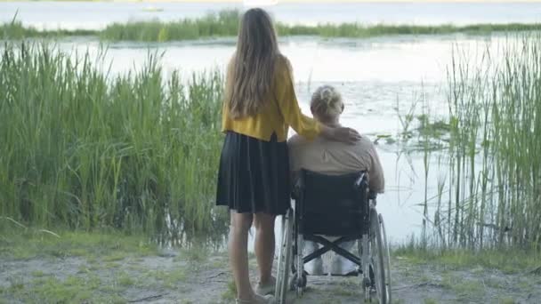 Baksidan beundrar det vita paret solnedgången på flodstranden. Ung kvinna och handikappad man på rullstol vilar utomhus på våren eller sommaren eller hösten dag. Kärlek och fritid. — Stockvideo