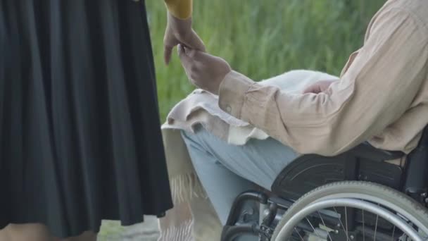 Nerozpoznatelná mladá žena hladící ruku postiženého muže sedícího na invalidním vozíku. Kavkazský pár si užívá rande venku v letní den. Postižený muž randí s přítelkyní. Koncept jednoty. — Stock video
