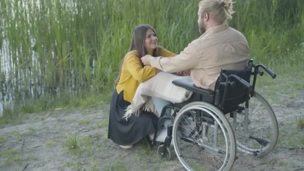 Paraplegický běloch sedící na invalidním vozíku a dotýkající se černých vlasů usměvavé ženy. Portrét šťastné veselé přítelkyně randící s postiženým přítelem na břehu řeky při západu slunce. Láska a romantika. — Stock video