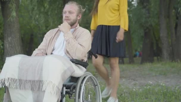 Неузнаваемая женщина приходит к мужчине, сидящему в инвалидном кресле и обнимающему парня. Портрет счастливого белого инвалида, встречающегося с девушкой в летнем парке вечером. — стоковое видео