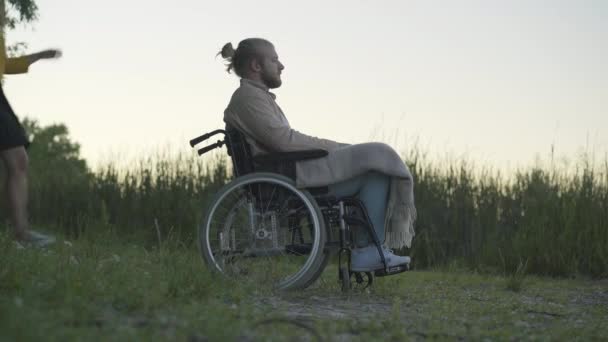 Vue de côté plan large de l'homme paralysé en fauteuil roulant reposant avec petite amie à l'extérieur au crépuscule. Portrait de heureux couple caucasien sortir ensemble au coucher du soleil. Pleine vie des personnes handicapées. — Video