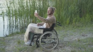 Tekerlekli sandalyedeki engelli adamın akıllı telefondaki selfie kamerasında görüntülü sohbet yoluyla konuşmasının yan görüntüsü. Gülümseyen Kafkasyalı erkek internette sohbet ediyor. Nehir kıyısında dinleniyor..