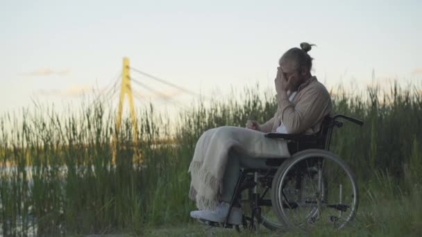 Vue latérale de l'homme handicapé déprimé assis en fauteuil roulant au coucher du soleil et regardant ailleurs. Portrait de paralysé désespéré caucasien invalide passer soirée solitaire sur la rive de la rivière. Concept Handicap. — Video