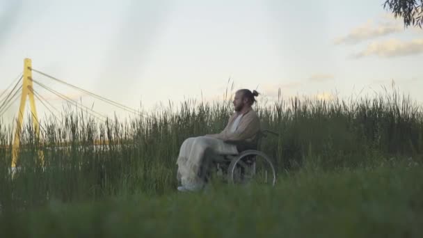 Široký záběr zamyšleného mladého handicapovaného muže na invalidním vozíku sedícího na břehu řeky a obdivujícího západ slunce. Boční pohled portrét pohledného postiženého bělocha invalidní těší letní večer venku. — Stock video