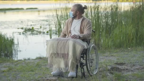 Široký portrét ochrnutého muže na invalidním vozíku s obličejovou maskou spočívajícím při západu slunce na břehu řeky. Pozitivně postižený kavkazský invalidní těší večer venku na Covid-19 uzamčení. — Stock video