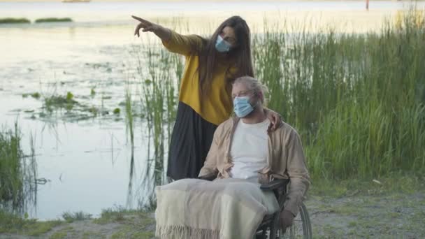 Portrait of confident young woman in face mask talking with paralyzed man in wheelchair. Wide shot of positive girlfriend dating with disabled boyfriend outdoors on Covid-19 pandemic quarantine. — Stock Video