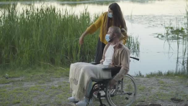 Boční pohled na bělošku v masce a invalidního muže na invalidním vozíku odpočívajícím na břehu řeky Covid-19. Mladá přítelkyně a zdravotně postižený přítel mluví venku při západu slunce. — Stock video