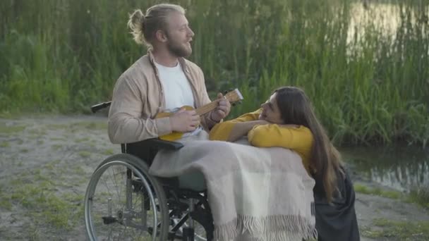 Kamera se blíží k úsměvu invalidní muž na invalidním vozíku hrát uklele pro krásnou mladou ženu. Portrét pozitivního kavkazského paralyzovaného přítele randění se šťastnou přítelkyní při západu slunce venku. — Stock video