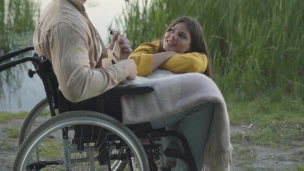 Mladá milující žena poslouchá paraplegického muže na invalidním vozíku hrajícího ukulele. Portrét krásné romantické kavkazské přítelkyně randění s handicapovaným přítelem při letním západu slunce venku. — Stock video