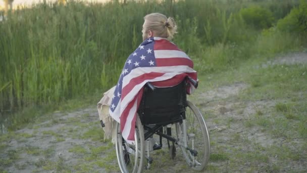 미국 국기에 싸여 휠체어에 앉아 생각하는 마비 된 남자의 뒷모습. 베테랑 남성이 여름 저녁을 혼자서 보내는 넓은 샷. 신체 장애와 신체적 상 해 개념. — 비디오