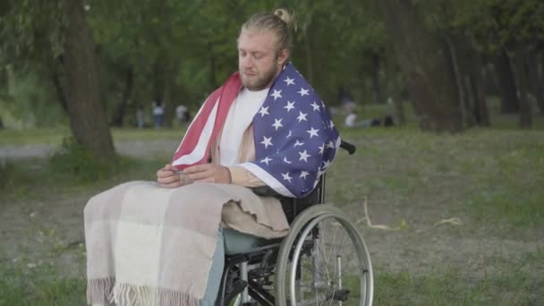 Ritratto di triste veterano paraplegico sulla sedia a rotelle che guarda strisce militari e sorride. La macchina fotografica si avvicina a soldato bello solitario che riposa in sera estiva in parco. Concetto di lesioni e disabilità. — Video Stock