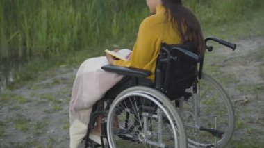 Dışarıda kitap okuyan engelli genç bir kadının arka açıdan bakışı. Tekerlekli sandalyedeki kendine güvenen beyaz kadın yazın edebiyatla parkta eğleniyor. Yaşam tarzı ve güven kavramı.