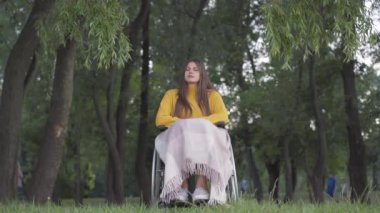 Parkta tekerlekli sandalyede oturan ve arka planda yürüyen insanlarla düşünen güzel beyaz kadın portresi. Engelli bayanın yaz akşamı tek başına dinlenme görüntüsü..
