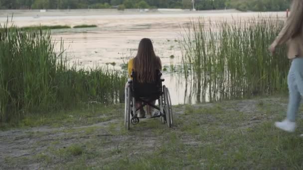 Vista posteriore di uomo amorevole venire alla donna disabile sulla sedia a rotelle e abbracciare la fidanzata. Felice ragazza paraplegica caucasica incontri con il fidanzato sulla riva del fiume al tramonto. L'amore non ha confini. — Video Stock