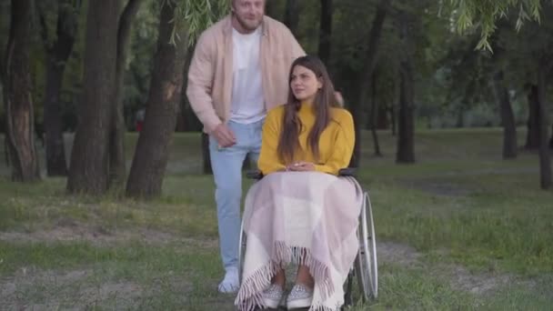 Szerokie ujęcie przystojnego kochającego mężczyzny przytulającego sparaliżowaną kobietę siedzącą na wózku inwalidzkim w parku. Portret młodej białej pary z plenerem w letni wieczór. Pojęcie miłości. — Wideo stockowe