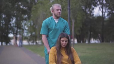 Akşam parkında tekerlekli sandalyede belden aşağısı felçli bir kadınla pozitif bir adamın portresi. Mutlu engelli bayan hemşireyle konuşuyor ya da gönüllü ve gülümsüyor. Sosyal hizmet kavramı.