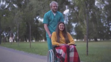 Beyaz adamın engelli bir kadınla yaz parkında yürürken ve ara sokakta dururken geniş açılı görüntüsü. Dışarıda felçli bir hastayla yürüyen gönüllü ya da hemşire portresi..