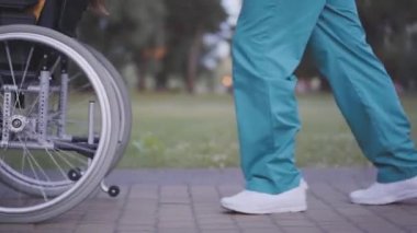 Tıbbi üniformalı, tanınmayan bir adamın, yaz parkındaki ara sokakta tekerlekli sandalyede yuvarlanışının yan görüntüsü. Bilinmeyen erkek hemşire ya da akşam vakti dışarıda engelli biriyle yürüyen gönüllü. Bakım kavramı