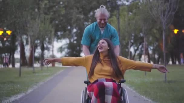 Retrato de una alegre mujer paralítica con las manos estiradas riendo como un enfermero corriendo rodando su silla de ruedas en el parque. Feliz señora discapacitada divirtiéndose con el cuidador caucásico y dejando. — Vídeo de stock