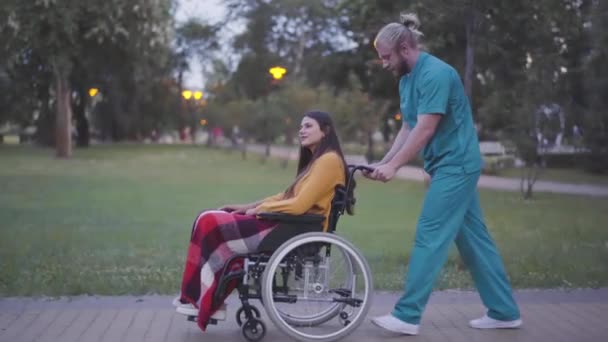 Vue latérale vue générale du fauteuil roulant roulant bénévole avec femme handicapée dans le parc d'été. Portrait d'un homme caucasien positif prenant soin d'une femme handicapée invalide. Bonne dame profiter de soirée. — Video