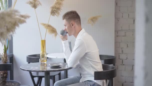 Vista lateral do empresário caucasiano confiante sentado à mesa no café e bebendo café. Jovem relaxado com camisa branca a fazer uma pausa na cantina. CEO masculino descansando dentro de casa. Conceito de negócio. — Vídeo de Stock
